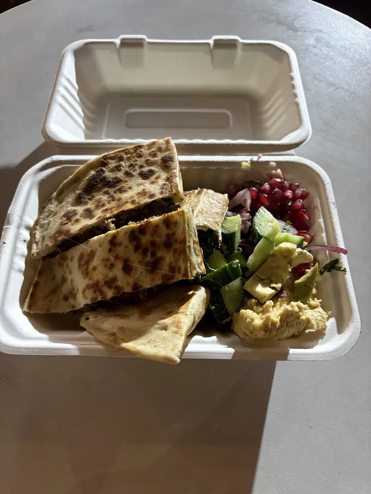 Open takeaway box with slices of gözleme, cucumber and pomegranate salad and hummus